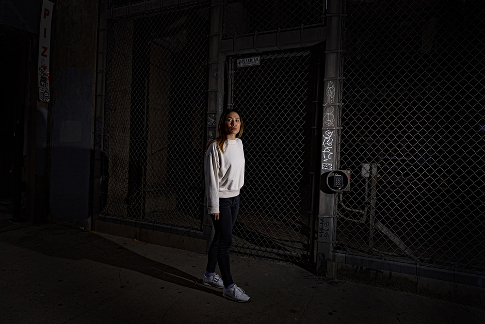  Young Asian woman with shoulder-length dark brown hair wearing white sweatshirt and black skinny jeans and white sneakers looks directly at the camera sternly and stands in front of a dark chain-link fence on a desolate sidewalk at night.