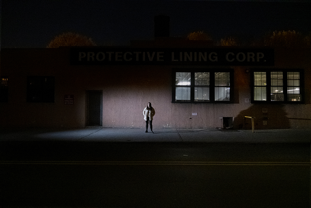 White woman with shoulder-length brown hair wearing cream-colored coat and pants stands across the street in front of a pink-colored industrial building along a desolate street at night..