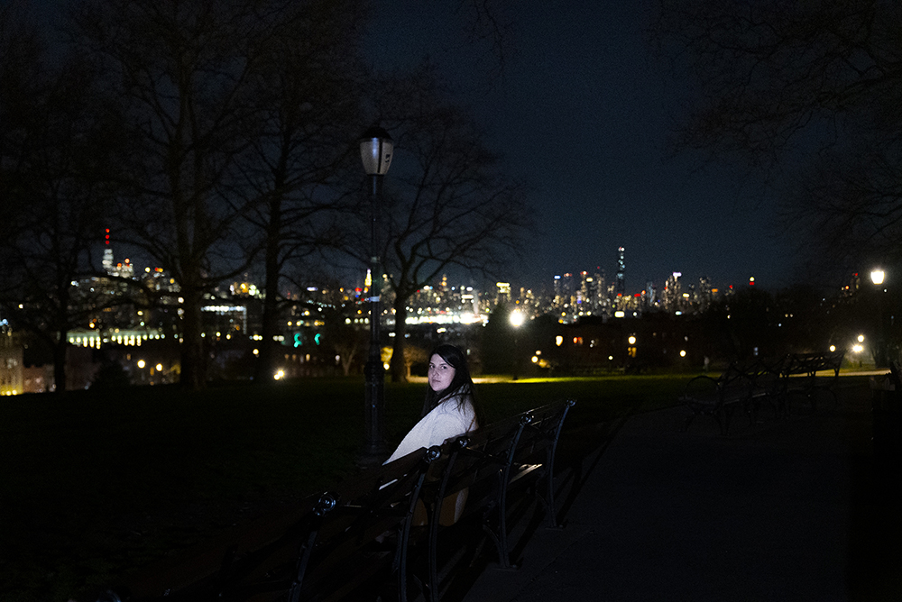  Caucasian woman with shoulder-length brown hair wearing cream-colored coat seen from behind, sits on a black bench in a dark city park and turns to look at the camera .