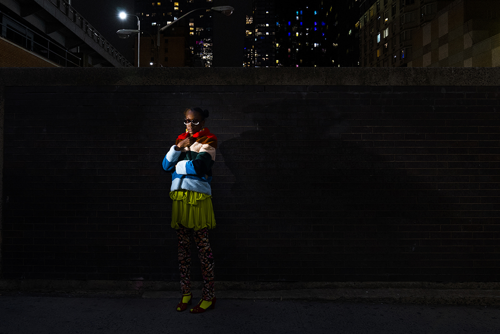 Black woman wearing brightly colored coat and lime-green dress clutches her jacket and looks sternly at camera while standing in front of brick wall in desolate NYC street